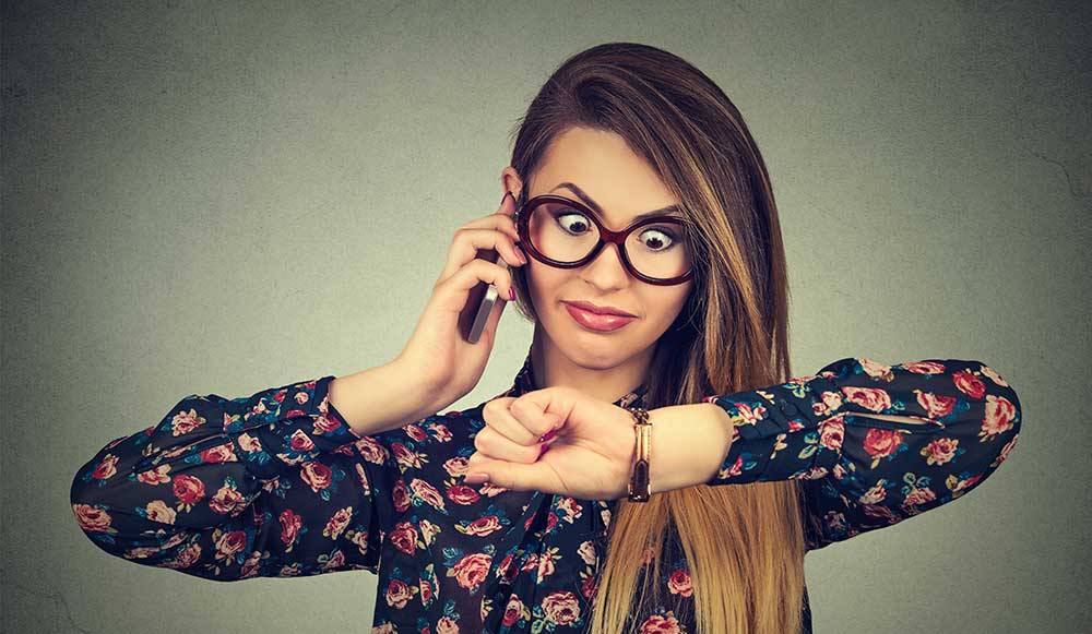 A lady in spectacles with a mobile phone to her ear and looking at her watch, indicating she is waiting for someone who is late to a meeting